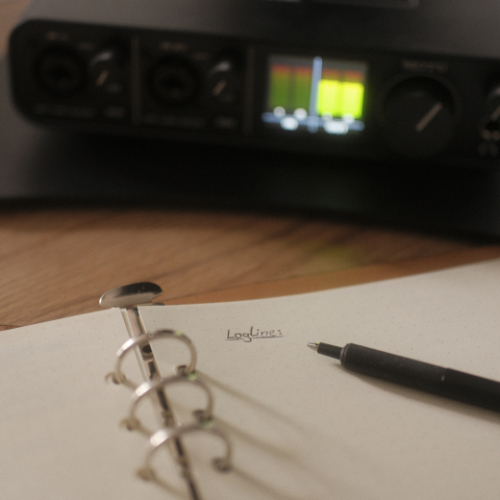 Paper with handwritten title 'Loglines' and a pen on a wooden desk with audio equipment in the background.