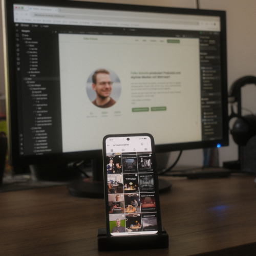 Smartphone displaying social media on a wooden desk, with a computer screen blurred in the background showing a webdesign software.