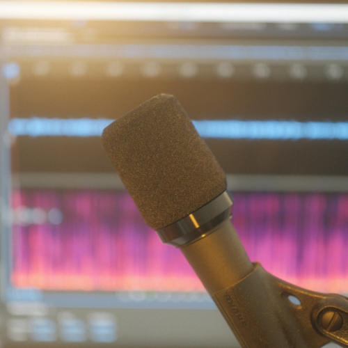 Black microphone with foam cover in front of a blurred digital audio waveform on a screen.